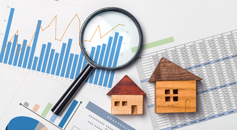 A magnifying glass and two small wooden houses placed on various financial charts and graphs.