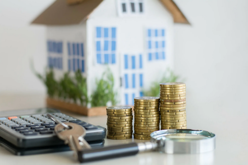 A miniature house, stacked coins, a calculator, and a magnifying glass are arranged on a table.