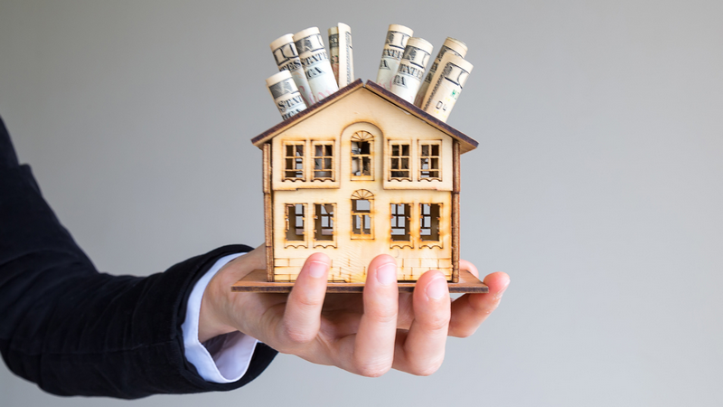 A hand holds a wooden house model filled with rolled-up dollar bills.