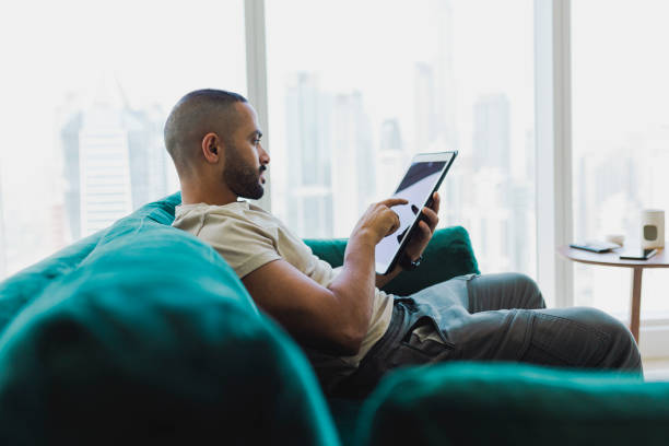 A man on a couch uses a tablet, researching how to buy property in Dubai.