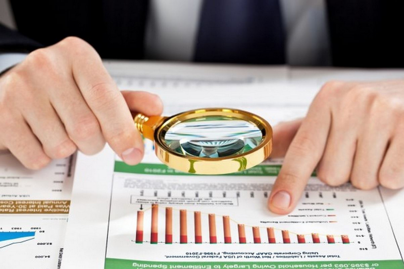 A person is using a magnifying glass to closely examine a financial document with charts and graphs.