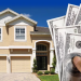 A hand holding a stack of 100-dollar bills in front of a large two-story house with a three-car garage.